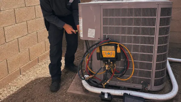 A technician with gauges hooked to an outdoor AC unit beside a block wall